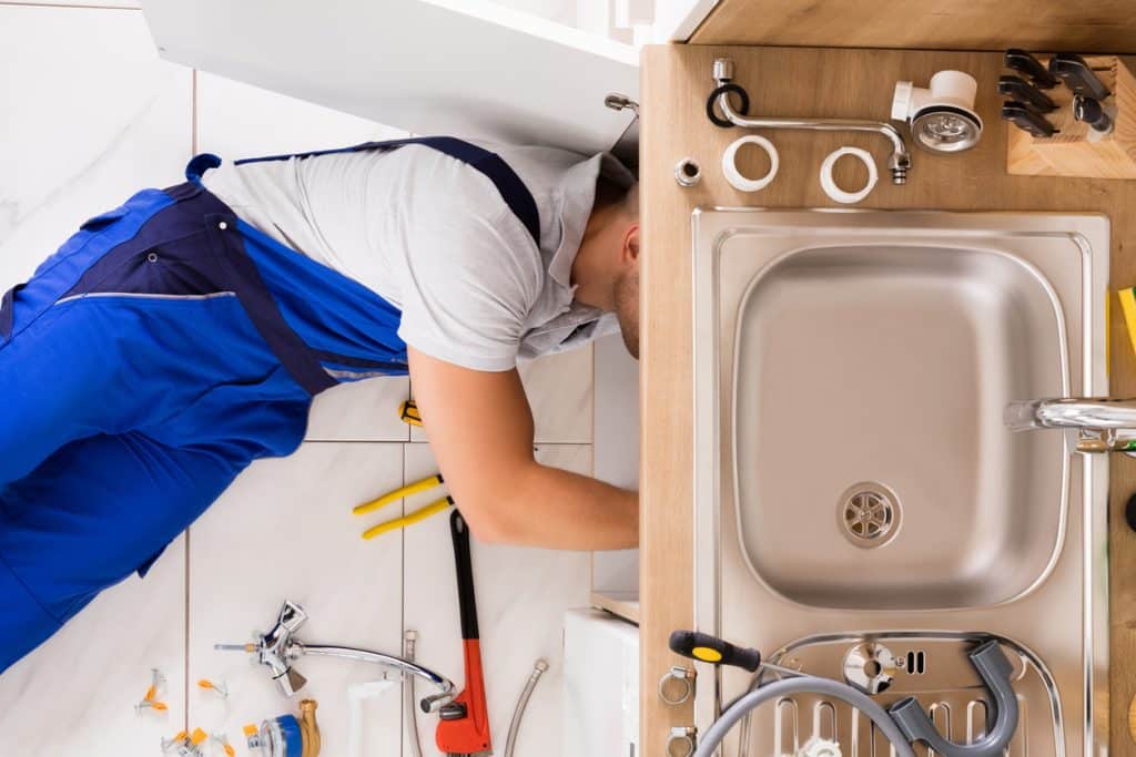 Plumber in blue overalls working under a kitchen sink, with plumbing tools and replacement fittings spread around the sink cabinet.